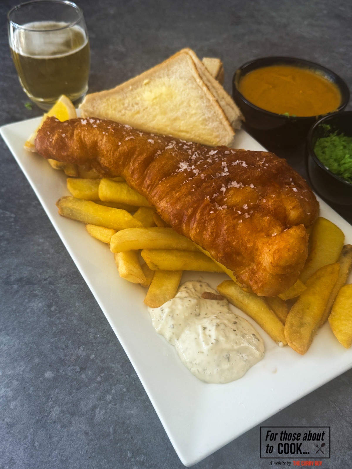 Beer batter fish and chips on a plate with curry sauce, mushy peas and tartar sauce.